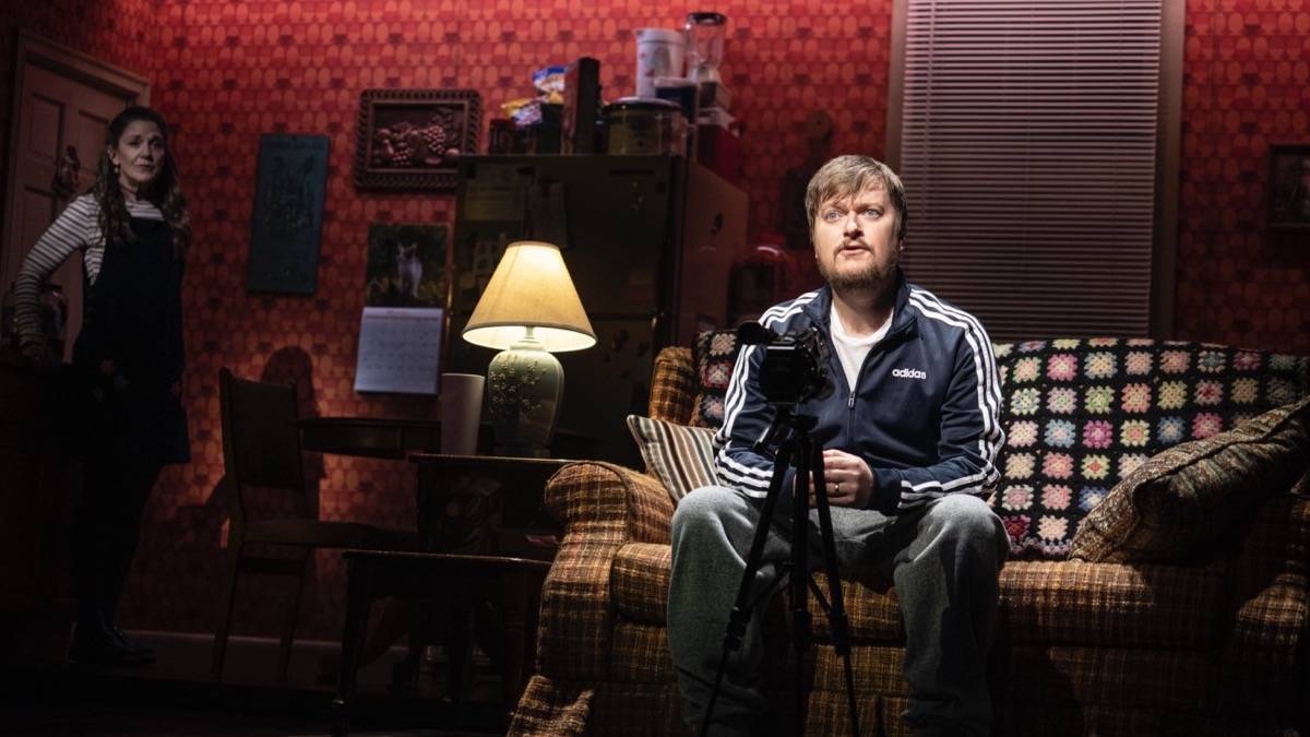 Man sitting on an old couch with a camcorder in front of him with a woman standing off to the side